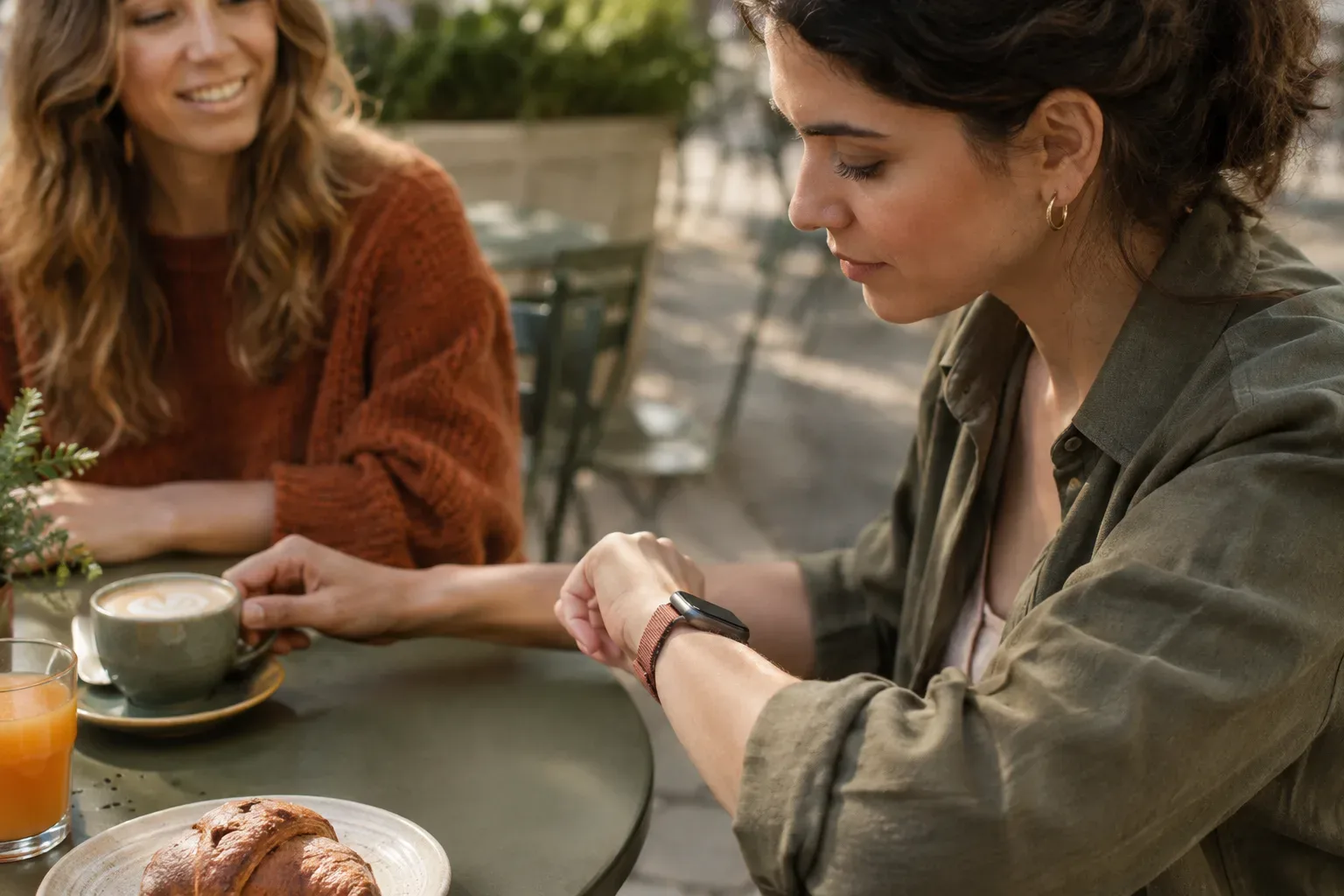 Baráti kávézás közben valaki egy terrakotta szövött szíjas Apple Watchra pillant, miközben nincs nála látható iPhone.